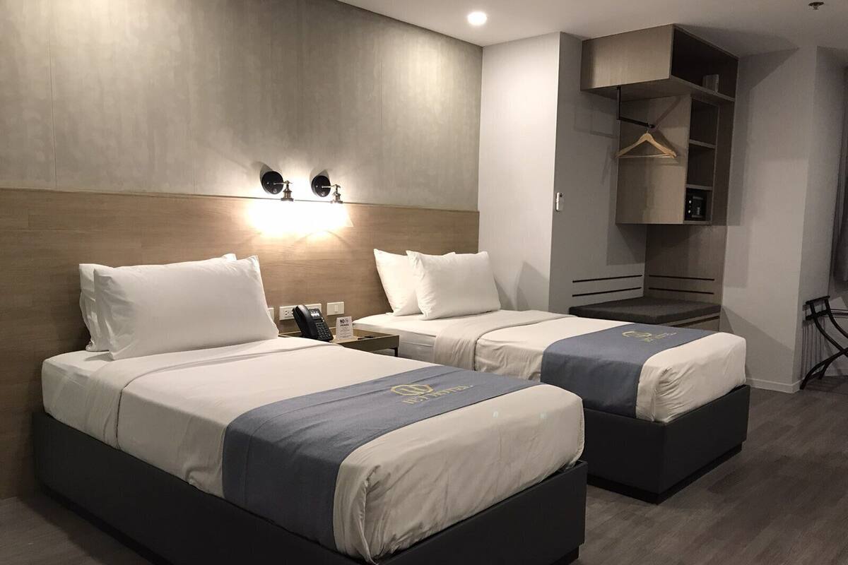 Quarto de hotel com duas camas, mesa de cabeceira com telefone, luminária na parede e guarda-roupa com cabideiro.