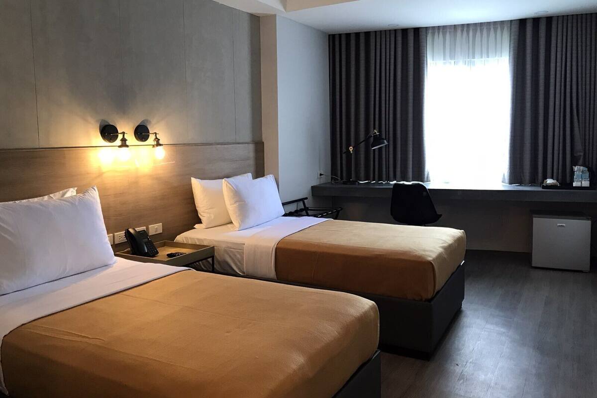 Quarto de hotel com duas camas, uma escrivaninha, uma cadeira, um abajur e um pequeno frigorífico.