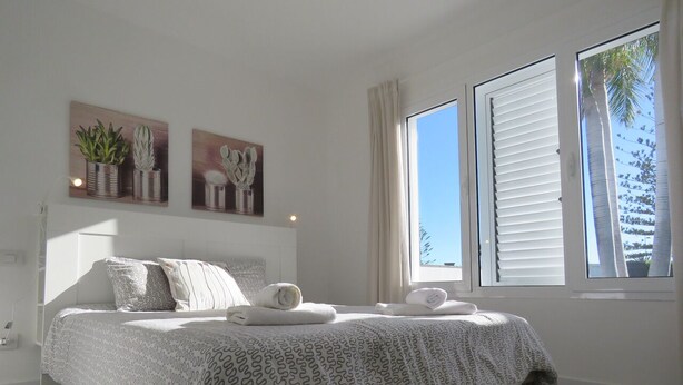 Een slaapkamer met een bed, kussens, een nachtkastje en een raam met jaloezieën.