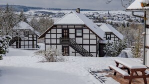 Une maison traditionnelle à colombages, avec un escalier menant à l’entrée, entourée d’arbres couverts de neige et offrant une vue sur une ville lointaine.