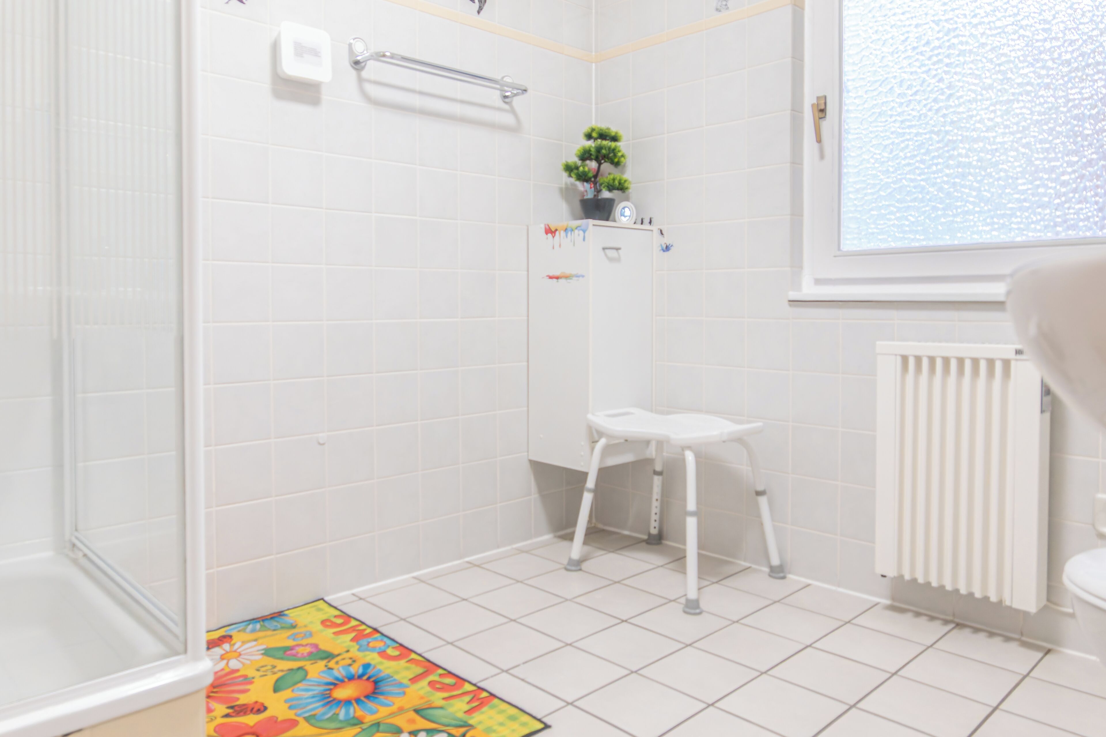 Ein Badezimmer mit weißen Fliesen, einer Dusche, einem WC, einem Heizkörper und einem Fenster.