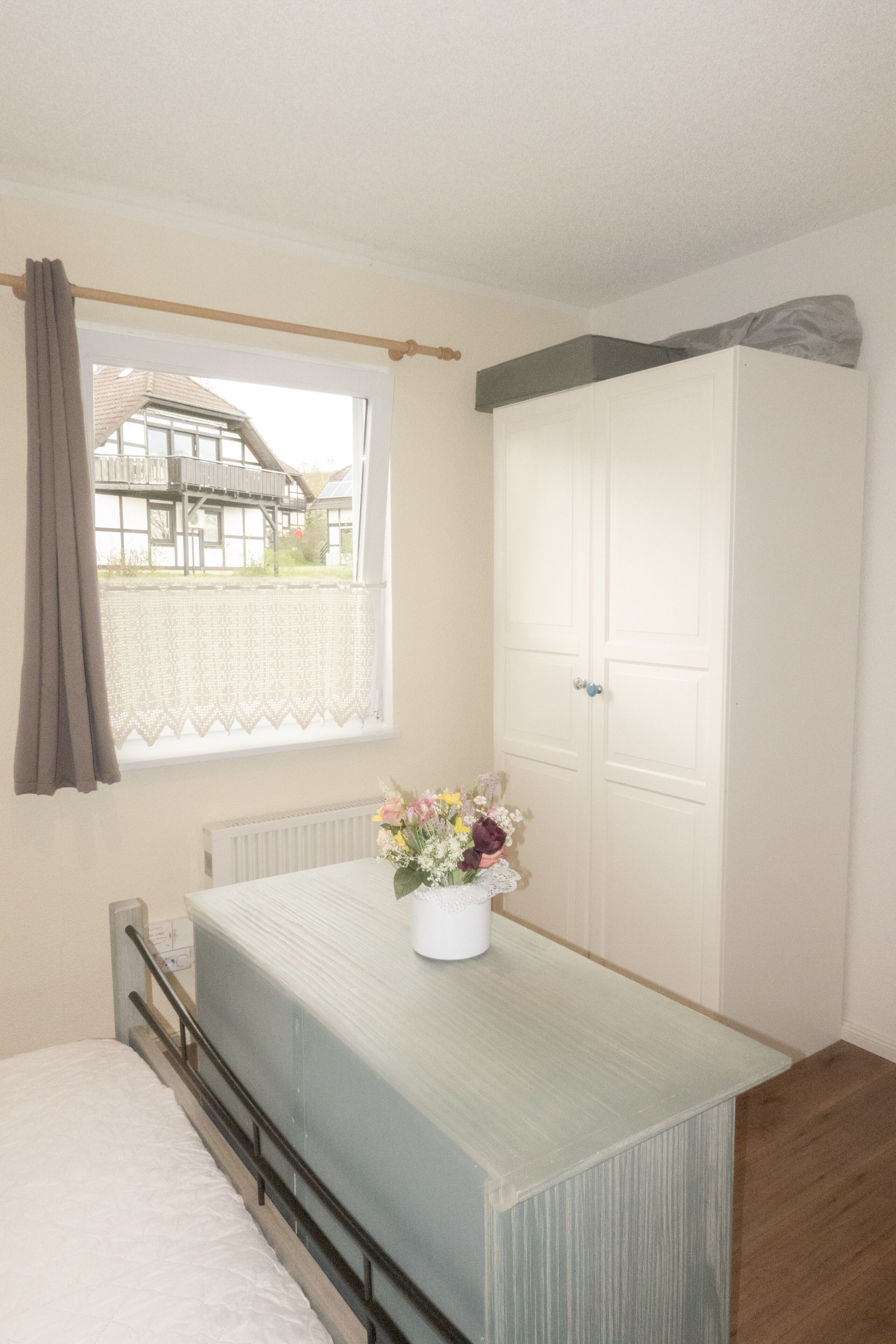 A bedroom with a bed, a window with a view of a house, a white wardrobe, and a vase with flowers on the bedside table.