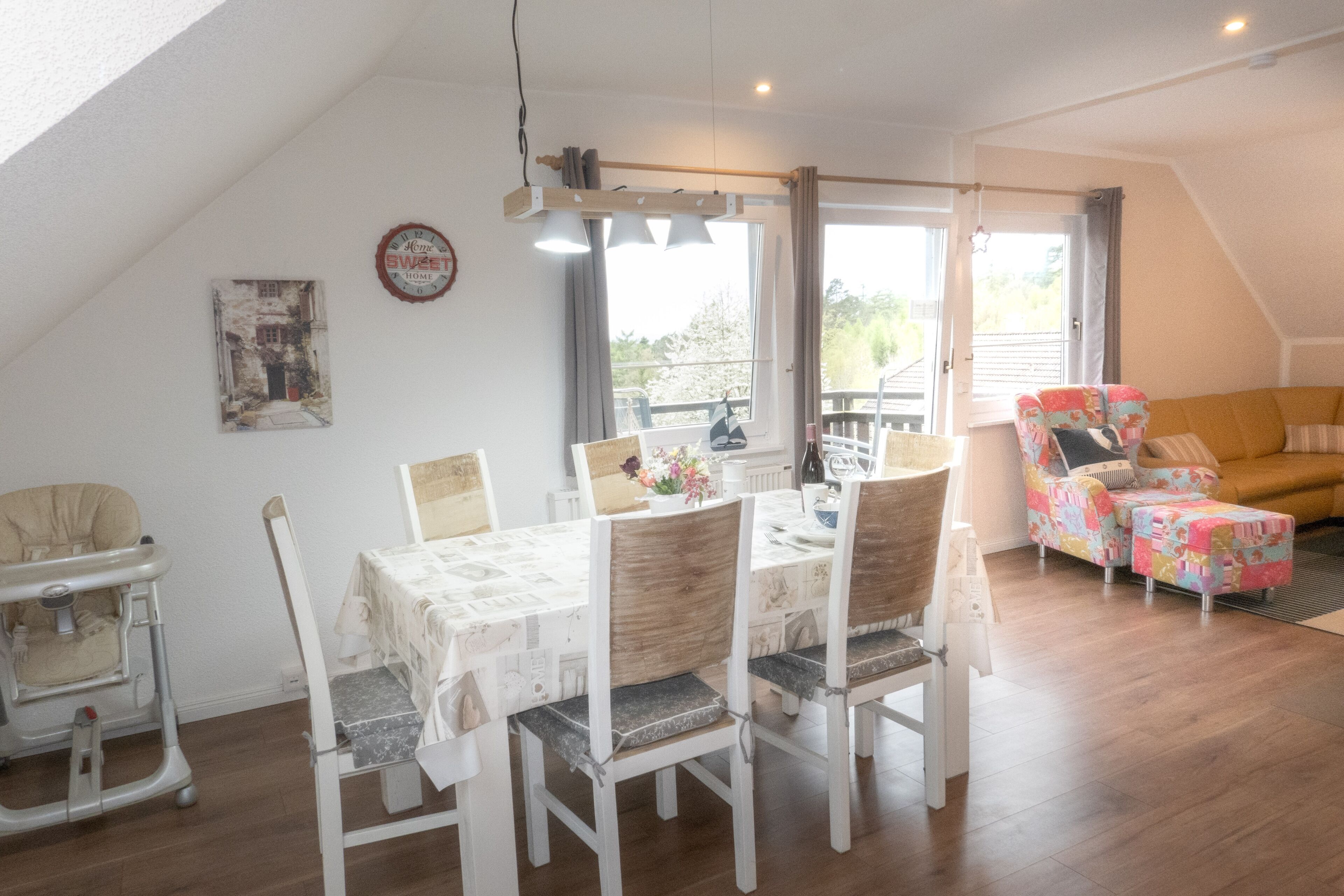 Un coin repas avec une table dressée pour quatre personnes, entourée de chaises. Il y a une chaise haute à gauche et une chaise colorée à droite.