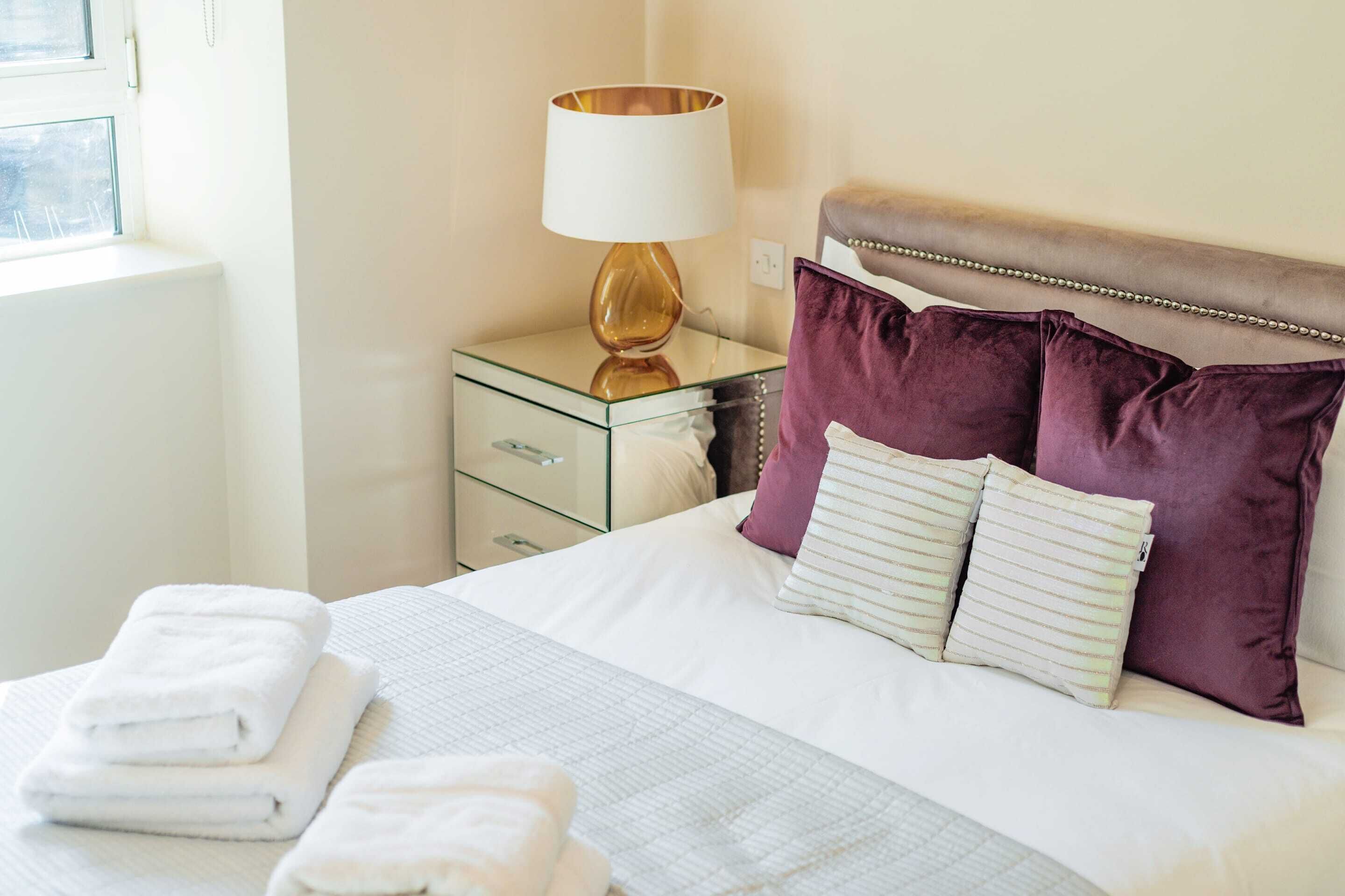A neatly made bed with white linens and striped pillows, a bedside lamp, and a glass nightstand with a drawer.