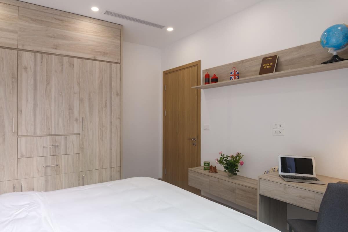 A bedroom with a bed, a desk with a laptop, a shelf with decorative items, and a wardrobe.