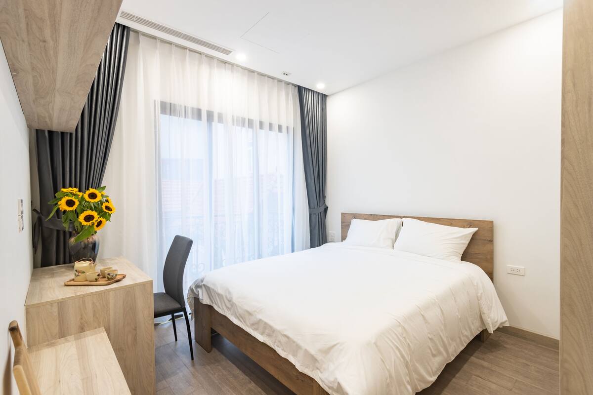 A modern hotel room with a wooden bed, a desk with a vase of sunflowers, and a chair.