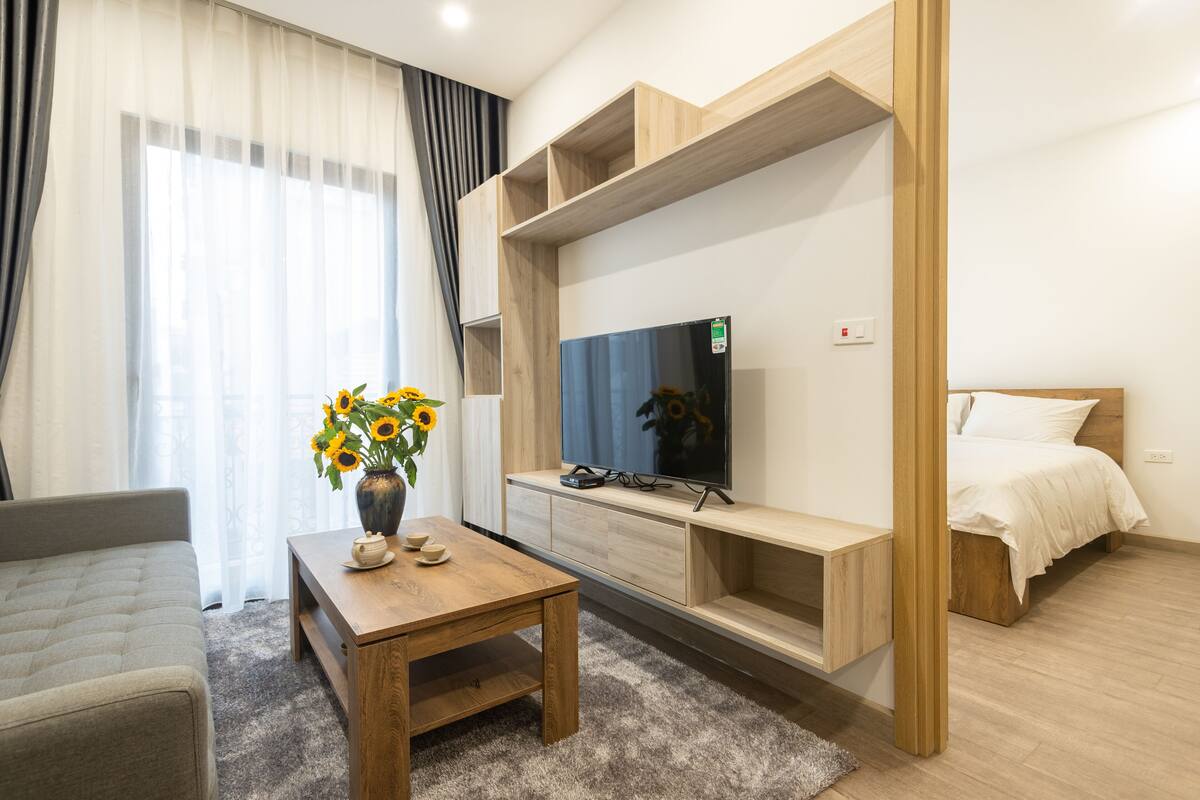 A modern hotel room with a bed, a TV, a wooden table with a vase of sunflowers, and a sofa.
