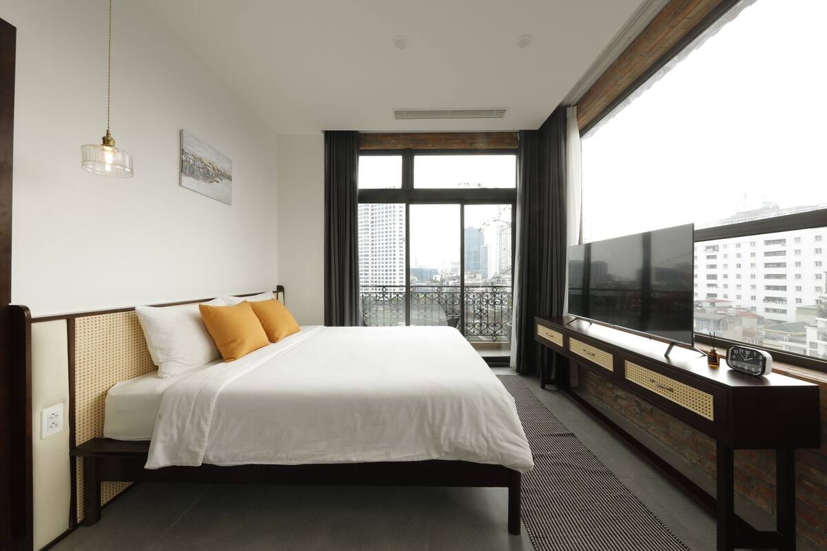 A hotel room with a bed, a TV, a window with a city view, and a hanging light fixture.