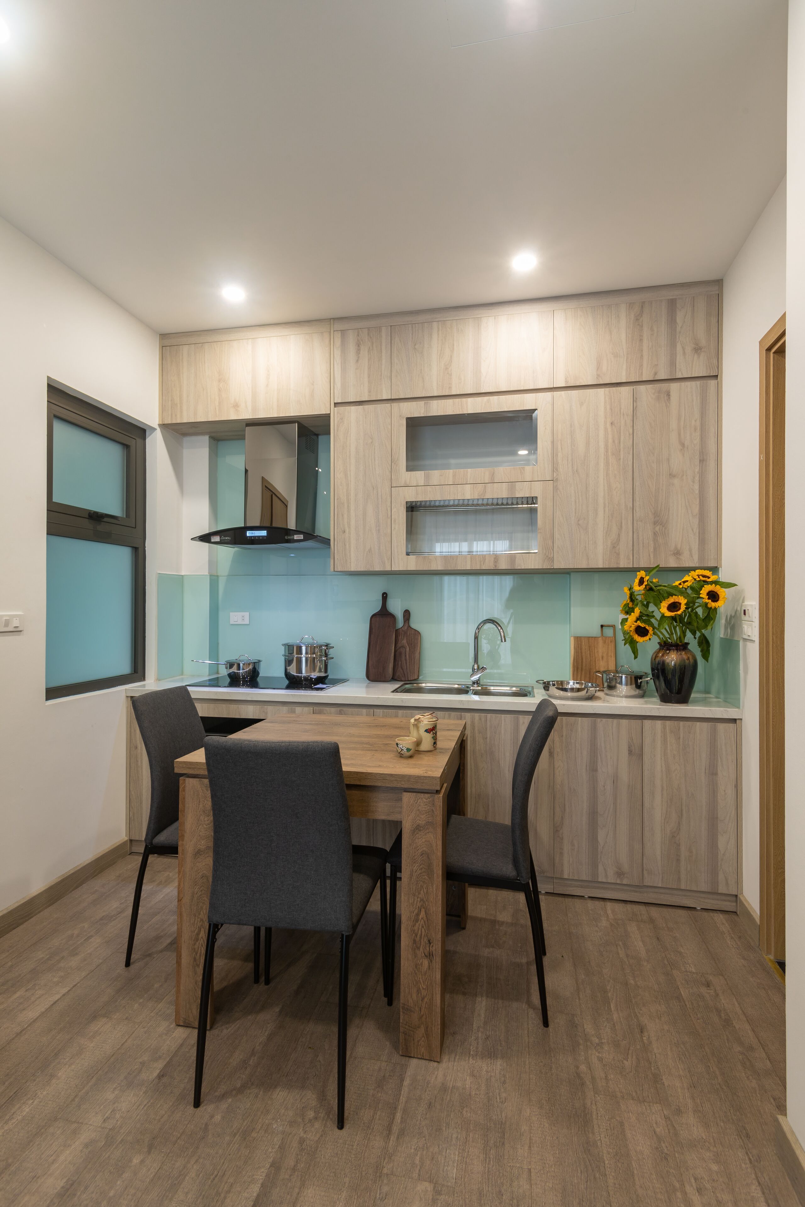 A modern kitchen with wooden cabinets, a dining table with chairs, and a vase of sunflowers on the counter.