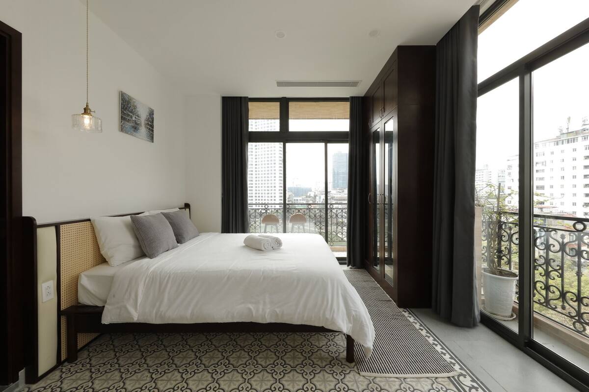 A modern bedroom with a large bed, a view of the city through a balcony, and a hanging light fixture.