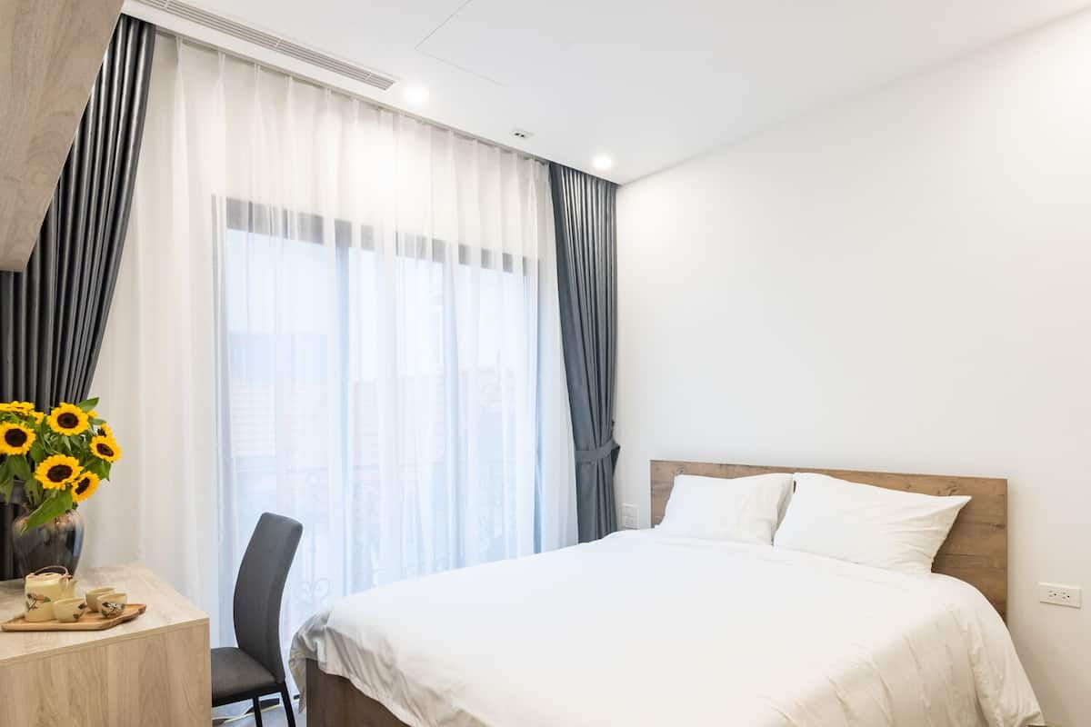 A hotel room with a bed, a desk with a chair, a vase with sunflowers, and a window with curtains.