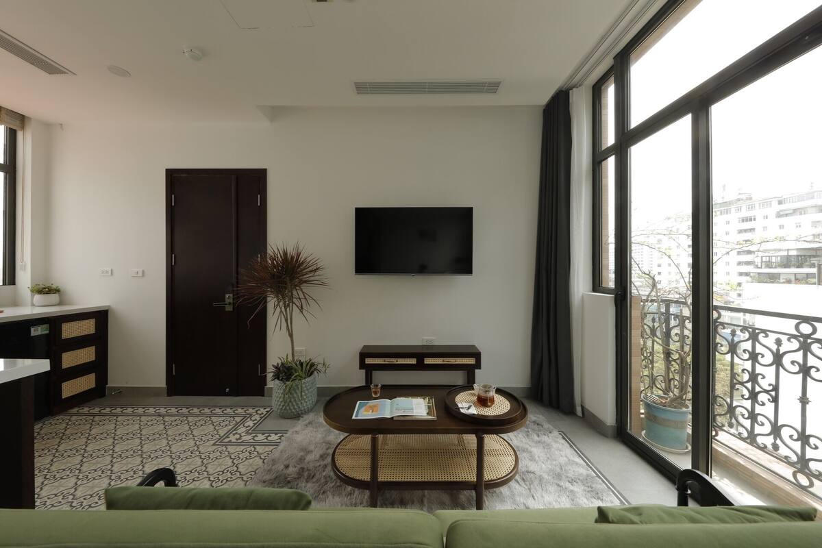 A modern living room with a sofa, a coffee table, and a TV. There is a large window with a view of buildings.