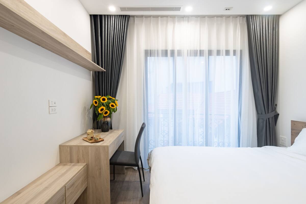 A modern hotel room with a bed, a desk with a chair, a small table with a vase of sunflowers, and a window with sheer curtains.