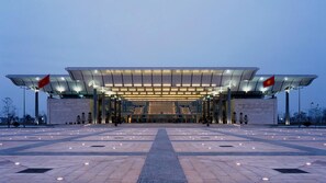 Un edificio moderno con una entrada amplia, dos banderas y un área pavimentada con luces.