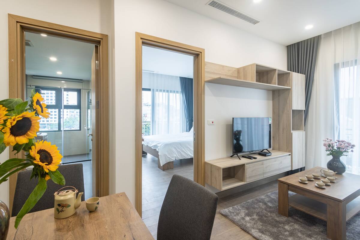 A modern hotel room with a wooden dining table, a sofa, a TV, and a bed with a white duvet.