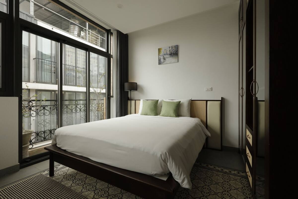 A hotel room with a large window, a bed with white bedding, a small painting on the wall, and a nightstand with a lamp.