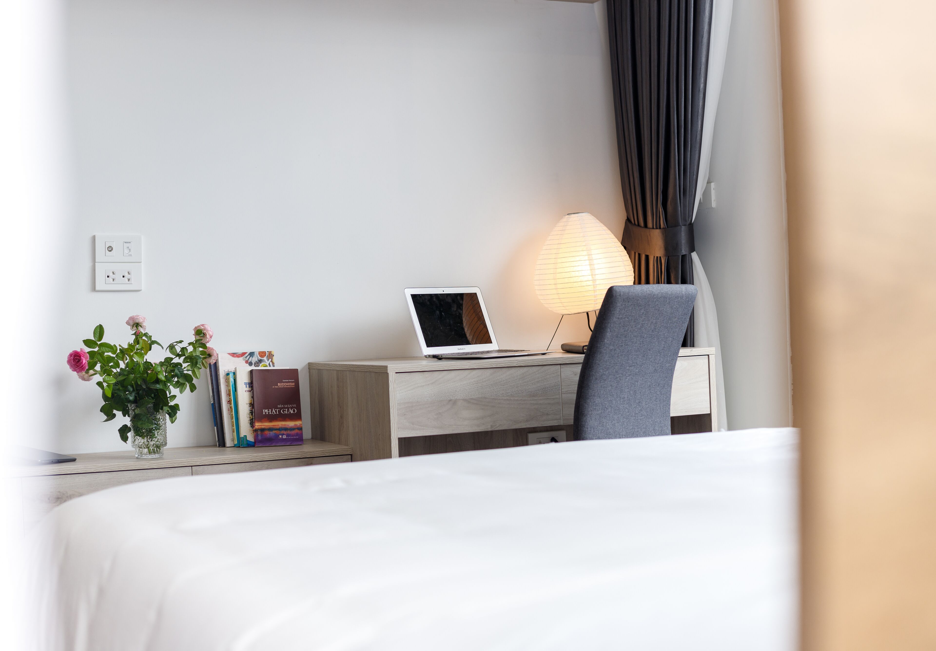 A hotel room with a bed, a desk with a laptop, a lamp, and a vase with flowers.