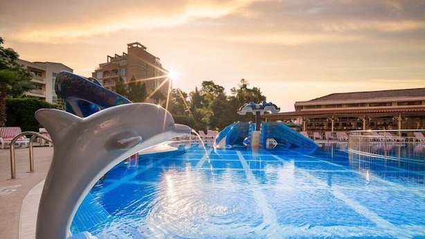 A swimming pool with a dolphin sculpture, a water slide, and a sun setting in the background. - Alba (Nessebar)