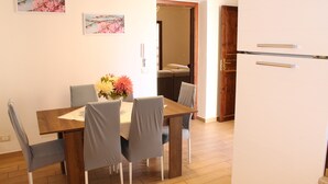 Un coin repas avec une table en bois, quatre chaises grises et un vase de fleurs. On aperçoit un réfrigérateur blanc et une porte en bois à l’arrière-plan.