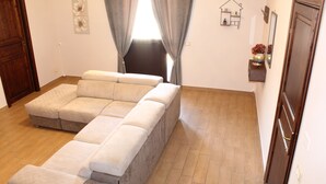 Un salon avec un canapé d’angle beige, un plancher en bois, une petite étagère ornée d’objets décoratifs et une porte.