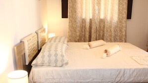 Un lit bien fait, recouvert d’une courtepointe blanche, avec deux serviettes enroulées, une lampe de chevet et un coussin à motifs posé sur une tête de lit en bois.