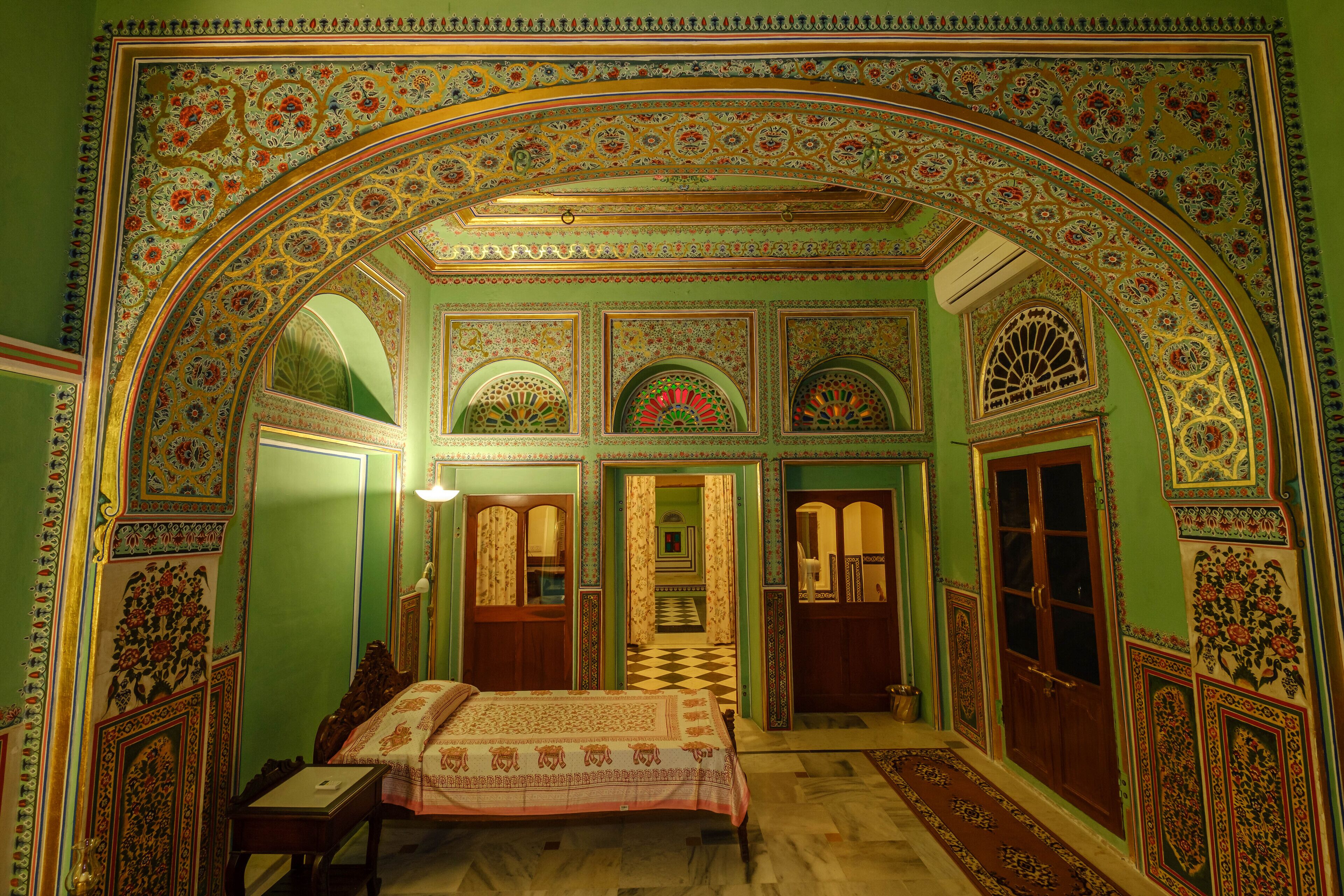 Une pièce comprenant un grand lit, un bureau et une chaise. Une porte donne accès à une autre pièce. Les murs sont décorés de motifs complexes.