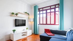 Una sala de estar moderna con un televisor de pantalla plana, un mueble blanco para entretenimiento, un sofá azul y una ventana con cortinas verde azulado.