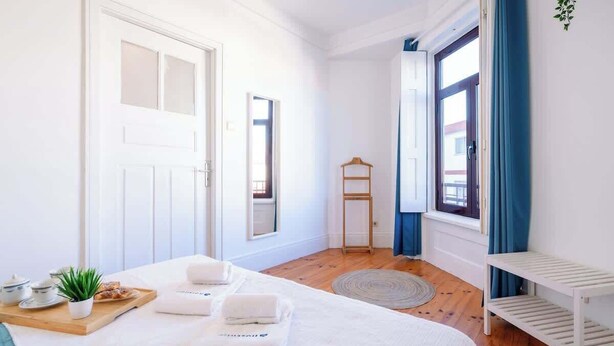 Una habitación luminosa y blanca con una cama, una mesita con una planta y una bandeja, y una ventana con cortinas azules.
