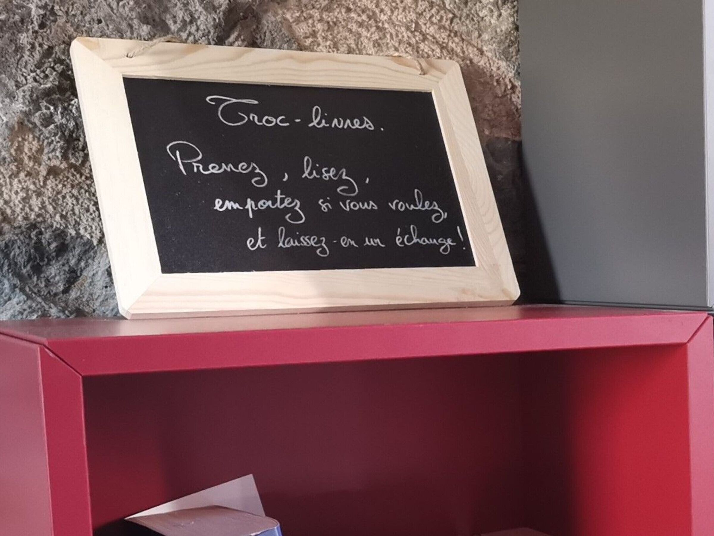 Een schoolbord met een boodschap in het Frans, in een houten lijst, geplaatst op een rode plank voor een stenen muur.