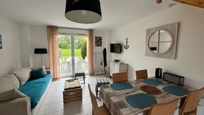 Un salon bien éclairé, avec un canapé, une table à manger et des chaises, un téléviseur et une grande fenêtre avec des rideaux.