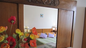 Hotelkamer met houten hoofdeinde, spiegel die de kamer reflecteert, bed met kussens, nachtkastje met lamp en vaas bloemen aan de linkerkant.