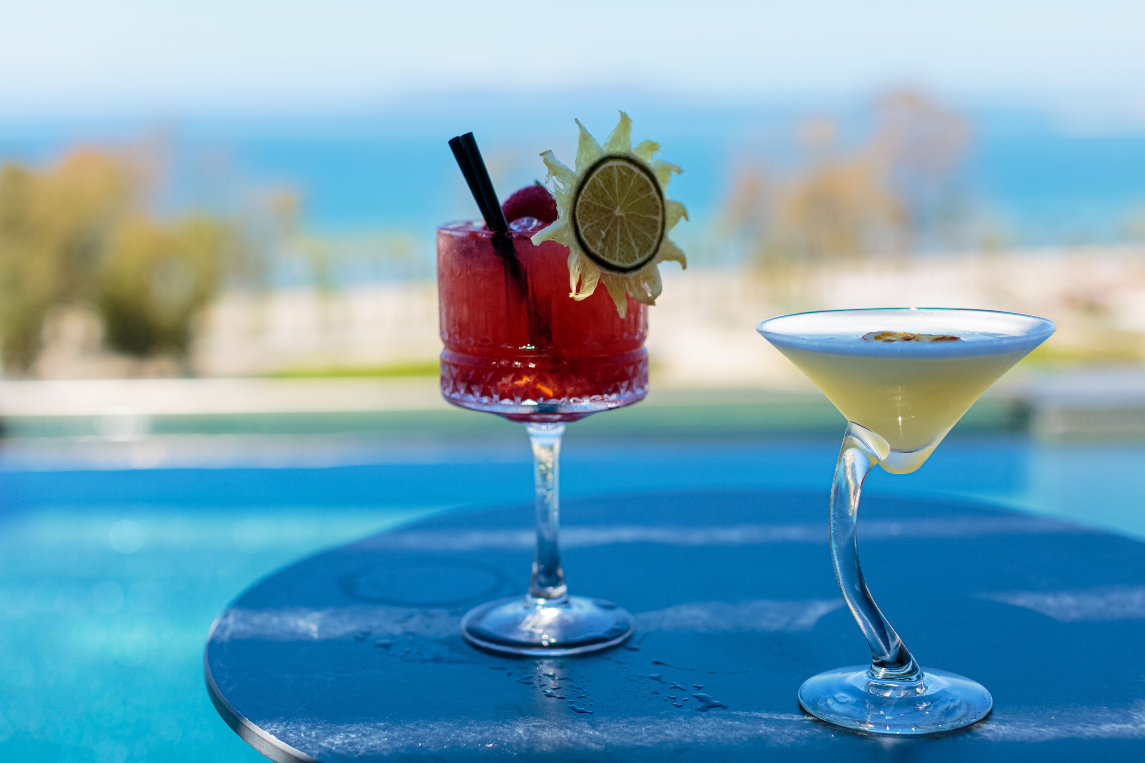 Two cocktails on a blue table by a pool with a blurred background.