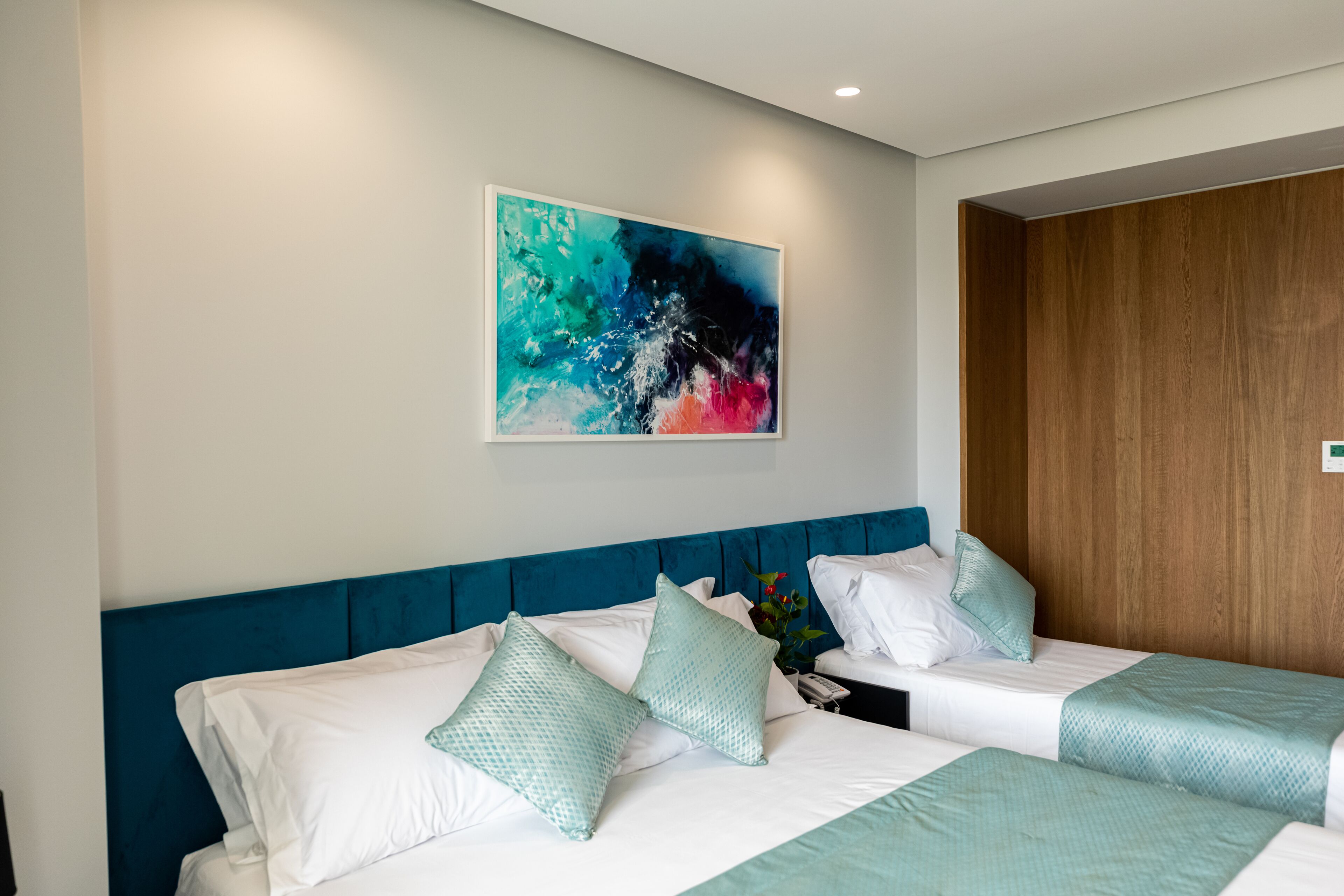 A hotel room with two beds, a blue headboard, white bedding, and a colorful abstract painting on the wall.