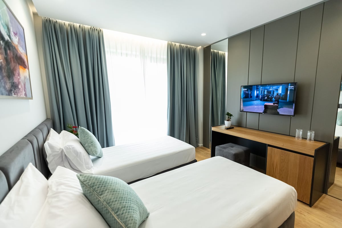 A hotel room with two beds, a desk, a TV, and a large window with curtains.