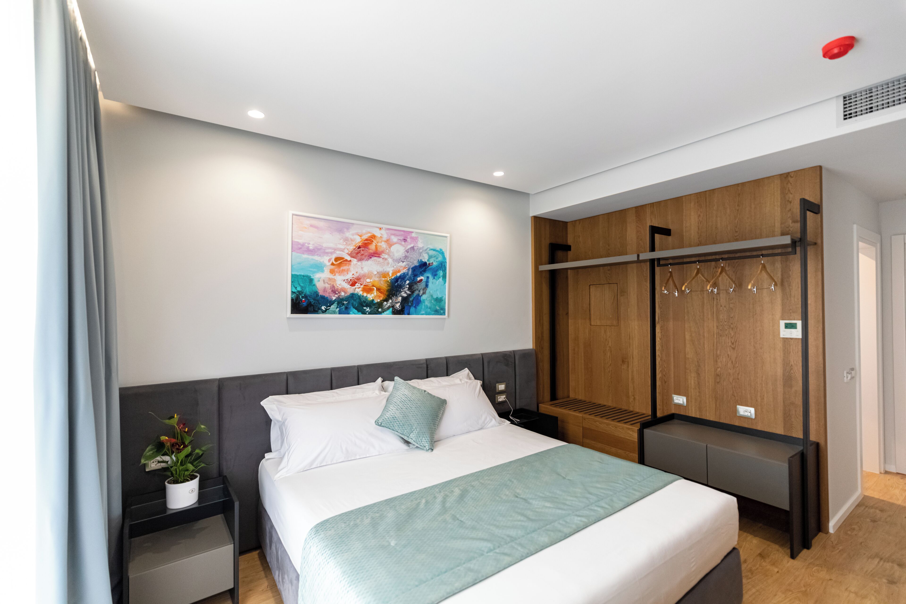 A modern hotel room with a bed, a bedside table with a plant, a wooden wardrobe, and a wall-mounted coat rack.