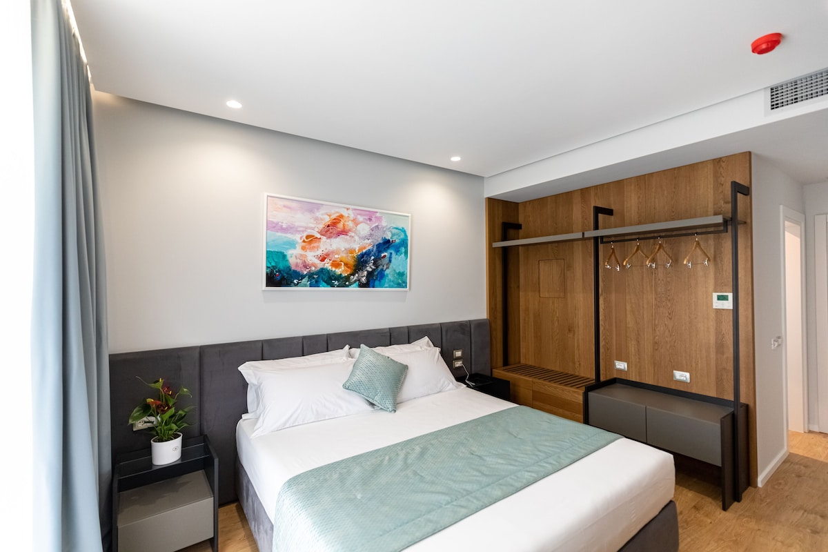 A modern hotel room with a bed, a nightstand, a wall-mounted coat rack, and a painting.