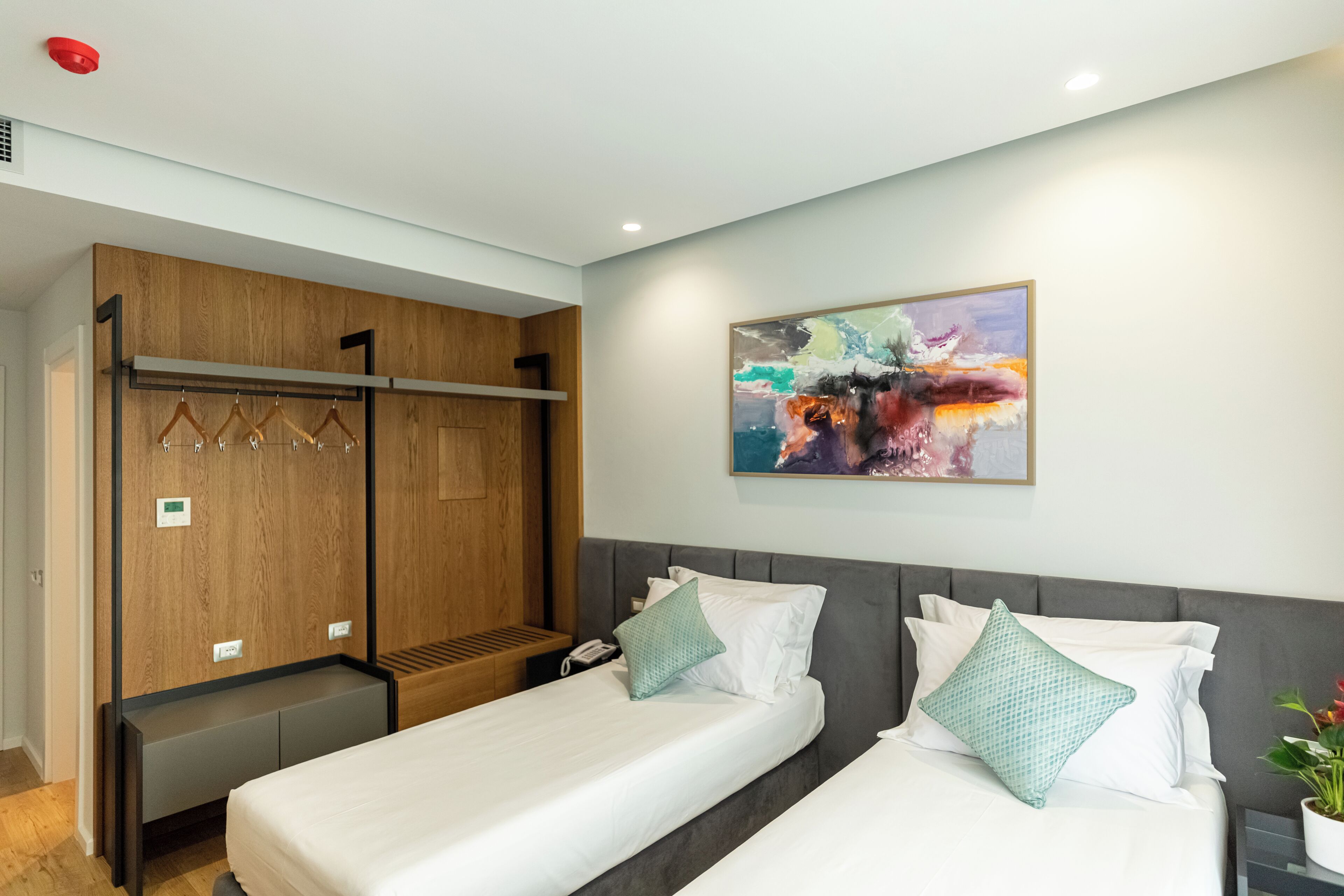 A hotel room with two beds, a wardrobe, a painting on the wall, and a potted plant.