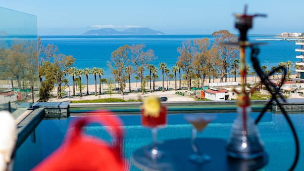 A poolside view with a hookah, a glass of drink, and a beach with palm trees and a distant mountain.