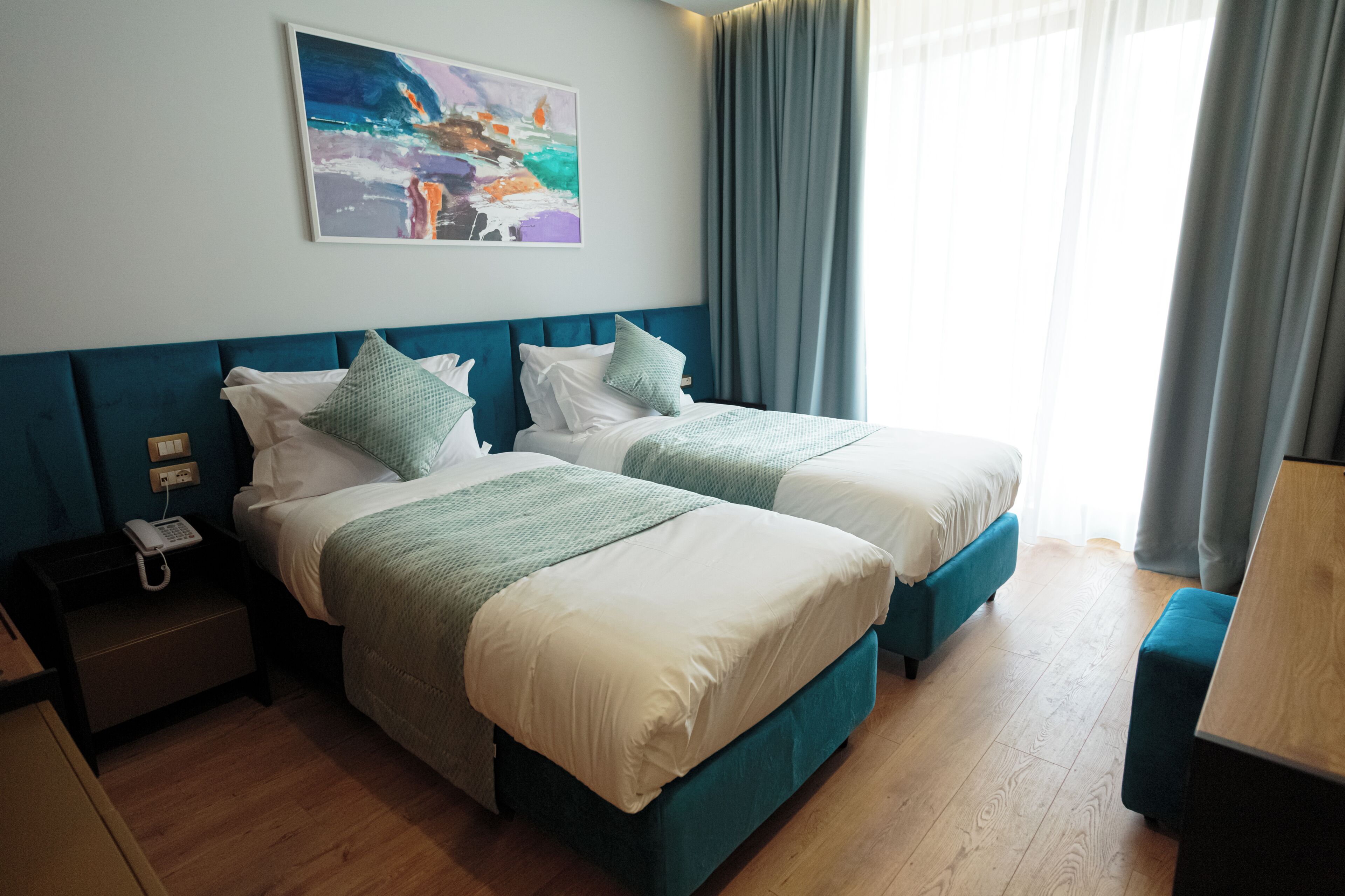 A hotel room with two beds, a nightstand with a phone, a painting on the wall, and a window with curtains.