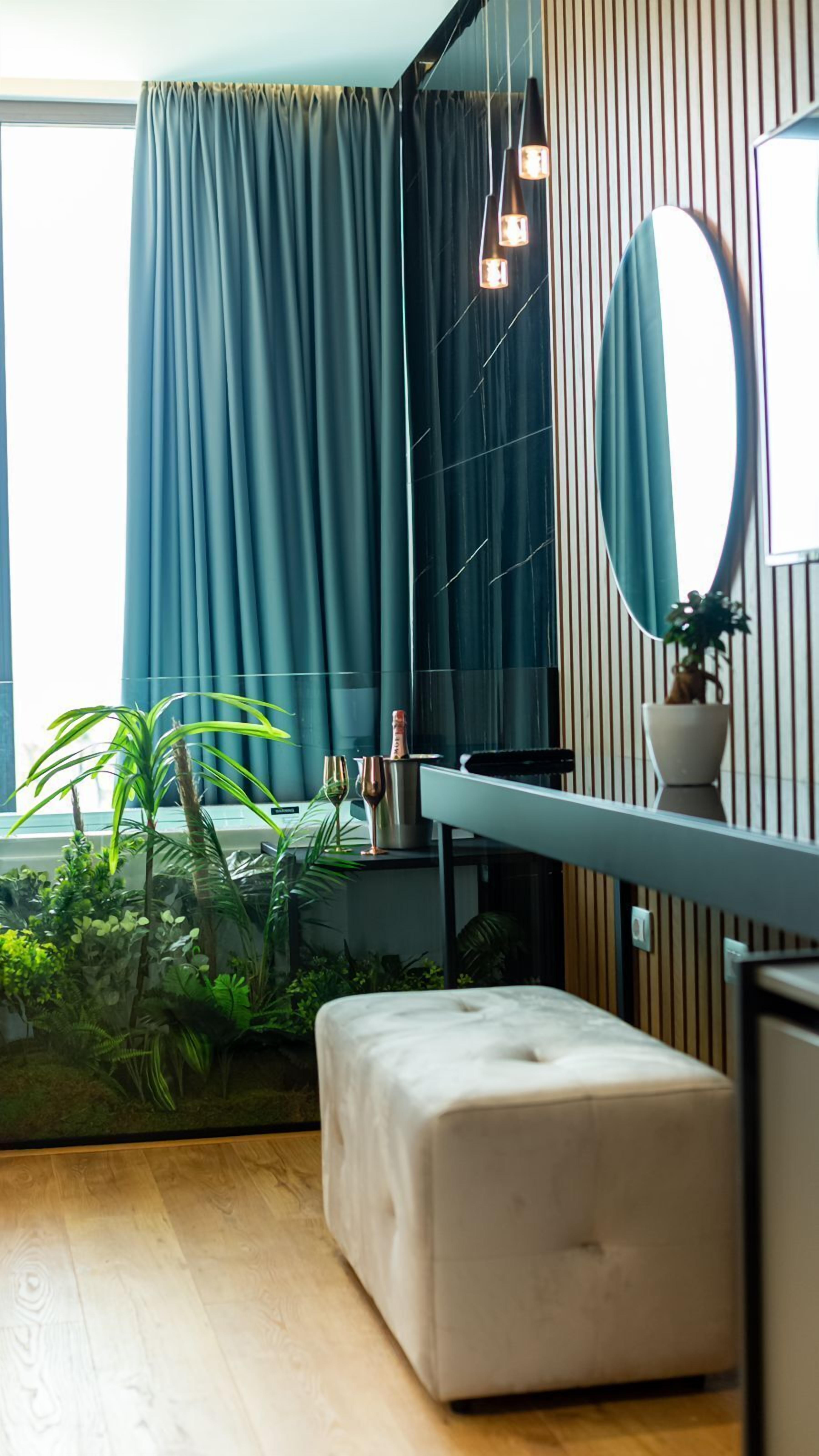 Una habitación moderna con piso de madera, un reposapiés blanco, un espejo, una planta en maceta y una ventana con cortinas.