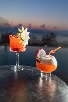Twee cocktails op een tafel met uitzicht op de oceaan en een zonsondergang op de achtergrond.