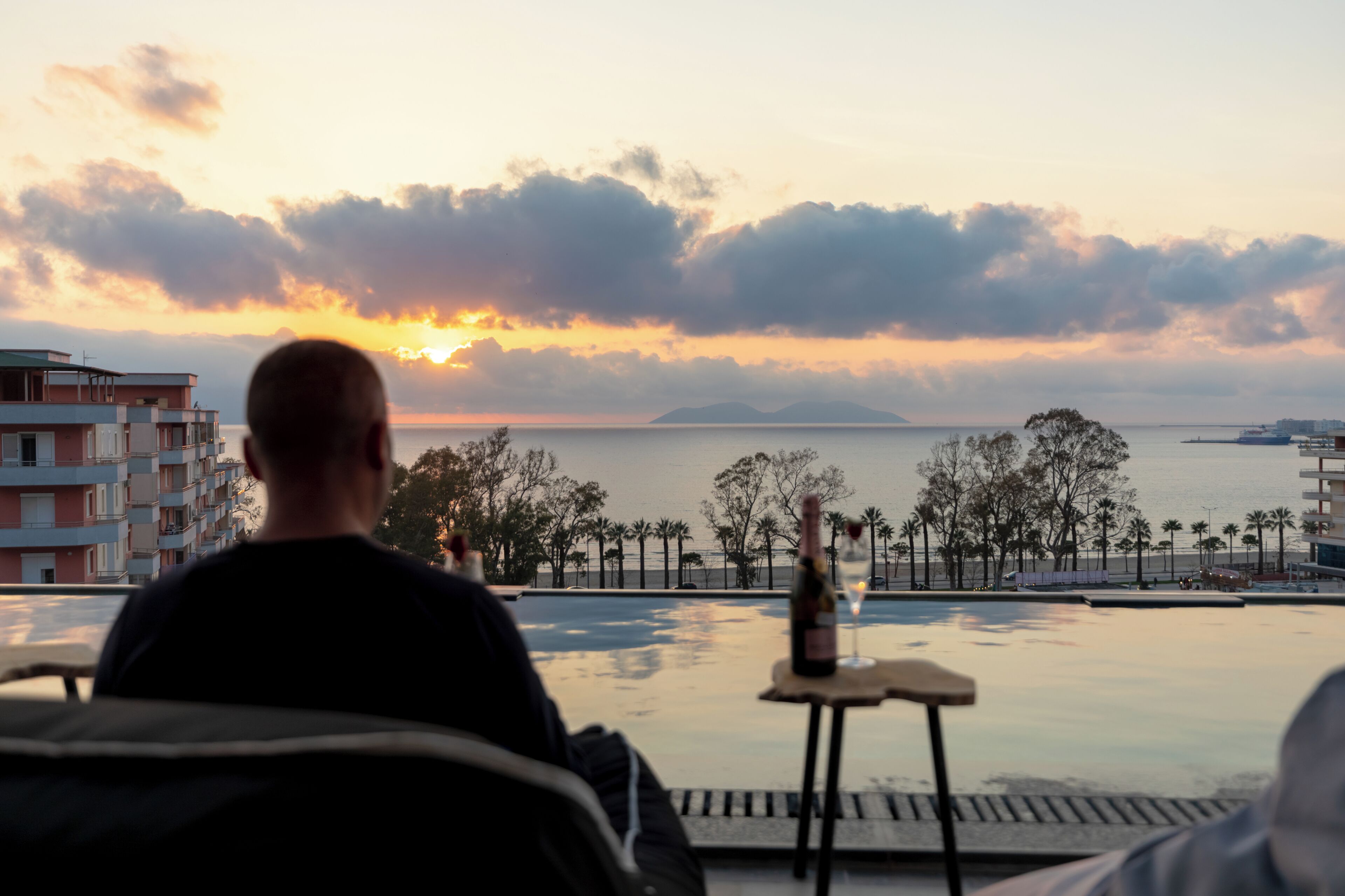 Une personne assise au bord d’une piscine à débordement, avec une vue sur le coucher de soleil, une bouteille de vin et une petite table.