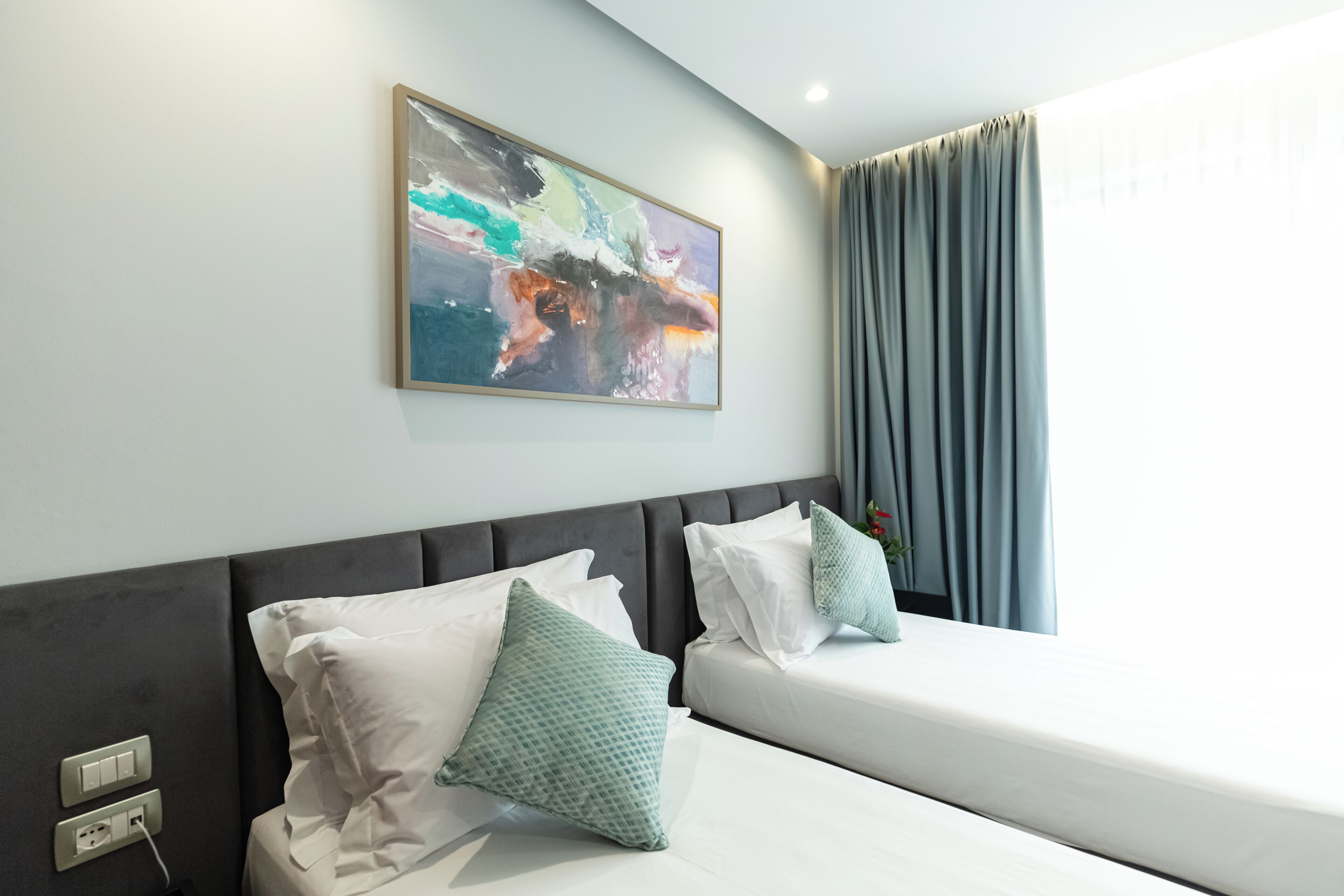 Habitación de hotel con diseño moderno, que incluye dos camas con ropa blanca y almohadas turquesa, un cuadro abstracto de gran tamaño en la pared, y una ventana con cortinas.