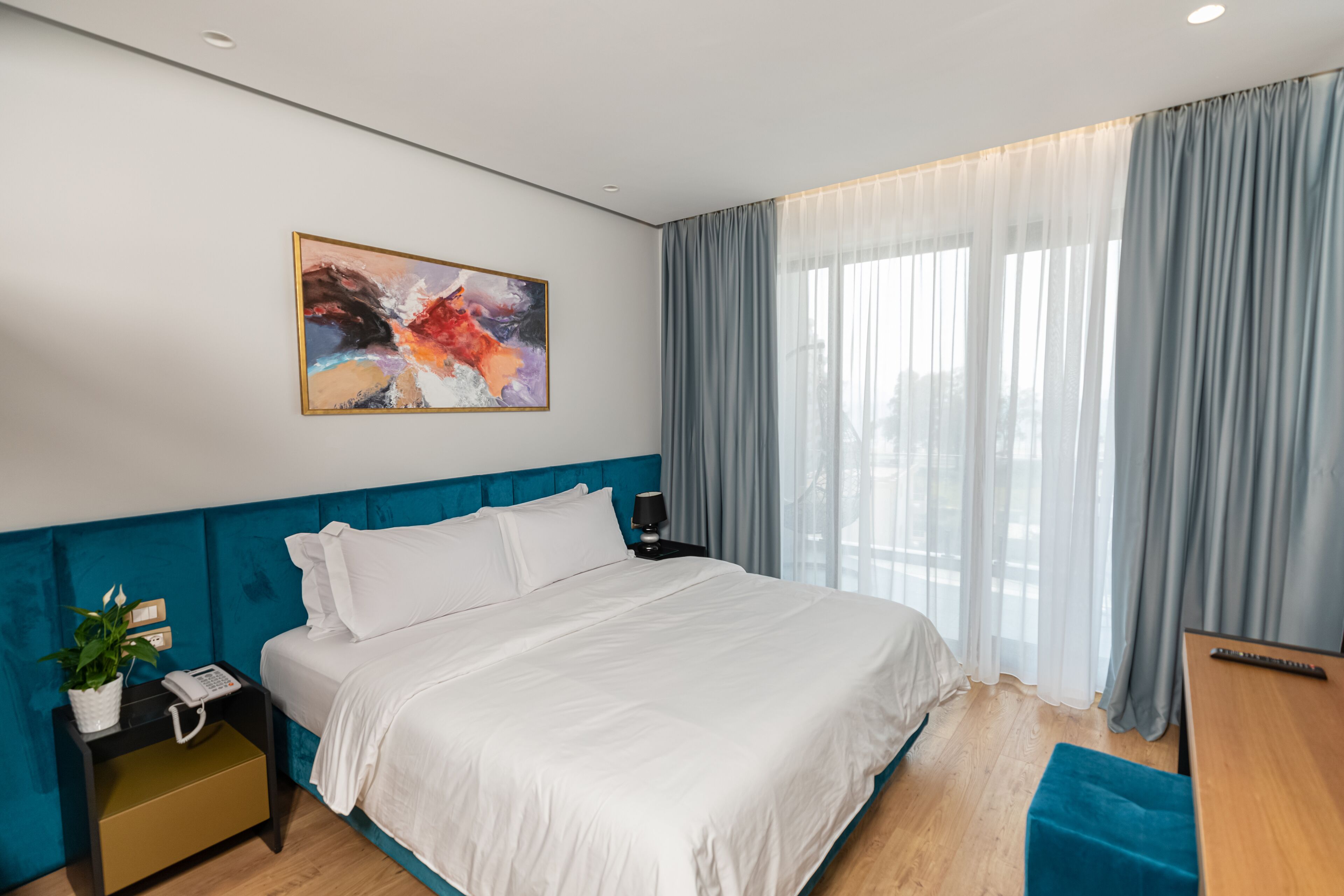 A modern hotel room with a large bed, a bedside table with a lamp, a phone, and a plant. There is a window with curtains and a painting on the wall.