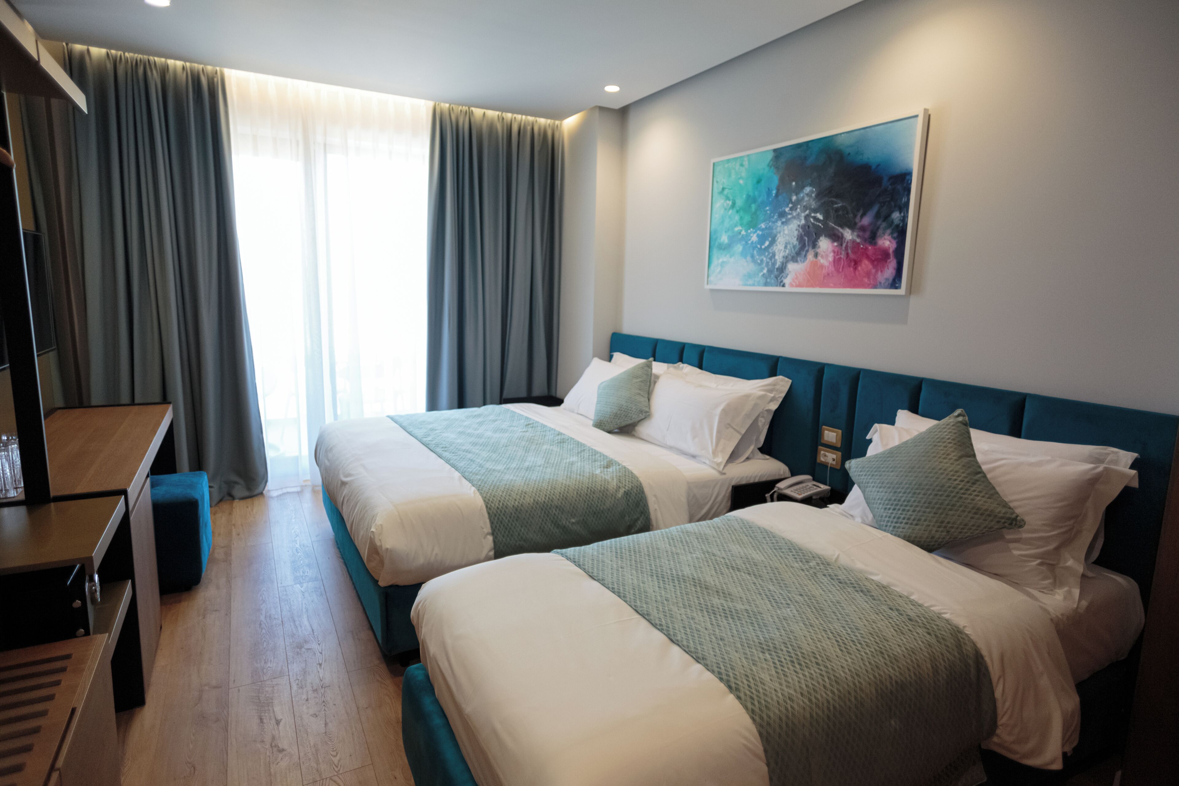 Habitación de hotel con dos camas, un escritorio, una silla, un cuadro en la pared y una ventana con cortinas.