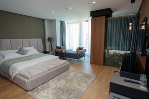 Moderni hotellihuone, jossa on suuri sänky, sohva, televisio ja ikkunasta avautuva näkymä vehreään maisemaan.