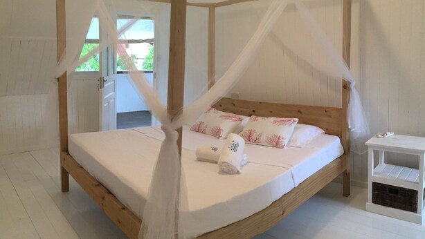 Uma cama de dossel com cortinas, roupa de cama branca e almofadas com padrão de folhas vermelhas, um criado-mudo de madeira e um banco branco.