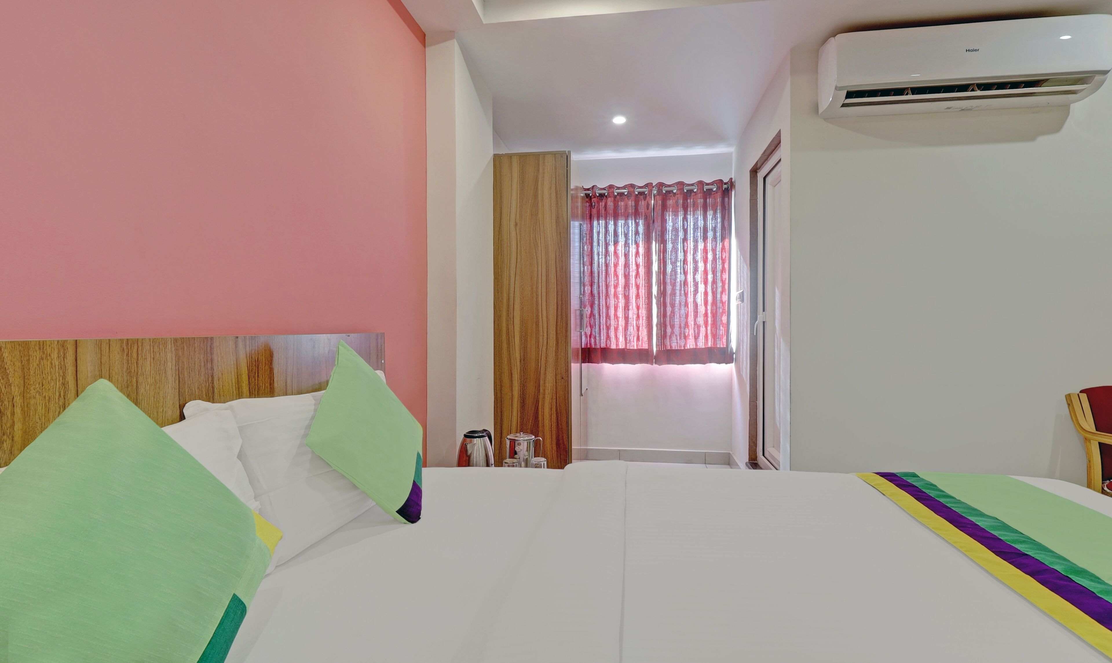 A hotel room with a bed, a wooden headboard, a window with red curtains, and an air conditioning unit.