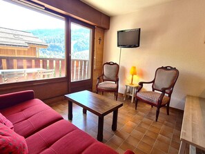 Una habitación con una mesa de madera, dos sillas, un sofá, un televisor montado en la pared y una ventana con vistas a un edificio y montañas.