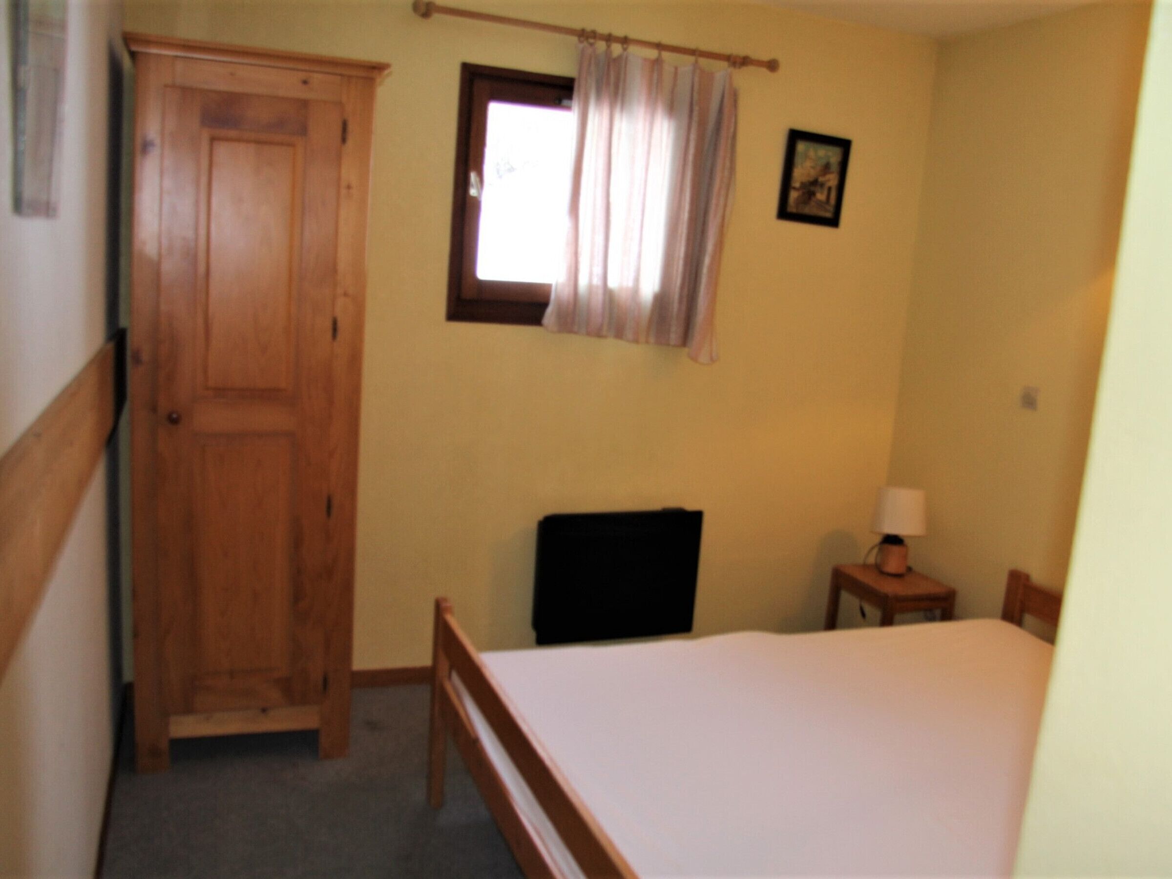 Een slaapkamer met een houten kledingkast, een enkel bed, een stoel, een nachtkastje met een lamp en een raam met gordijnen.